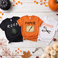 Three Halloween-themed t-shirts on a wooden surface with pumpkins and leaves.
