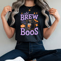 A woman wearing a black graphic t-shirt with a purple and orange 'cold BREW Boos' Halloween design, paired with dark blue jeans and white sneakers, sitting on a red stool.
