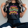 A woman wearing a black graphic t-shirt with a colorful 'FIRST HALLOWEEN MAMA' print, paired with dark blue jeans and white sneakers, sitting on a red stool against a plain light gray wall.
