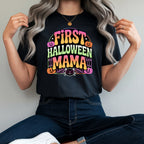 A woman wearing a black graphic t-shirt with a colorful 'FIRST HALLOWEEN MAMA' print, paired with dark blue jeans and white sneakers, sitting on a red stool against a plain light gray wall.
