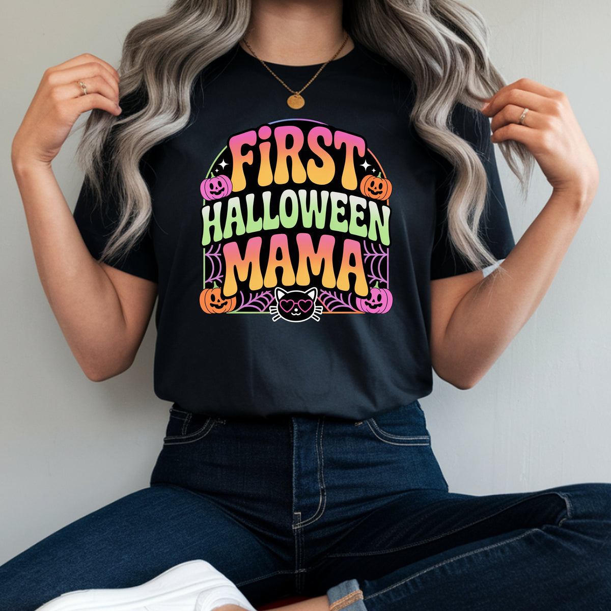 A woman wearing a black graphic t-shirt with a colorful 'FIRST HALLOWEEN MAMA' print, paired with dark blue jeans and white sneakers, sitting on a red stool against a plain light gray wall.