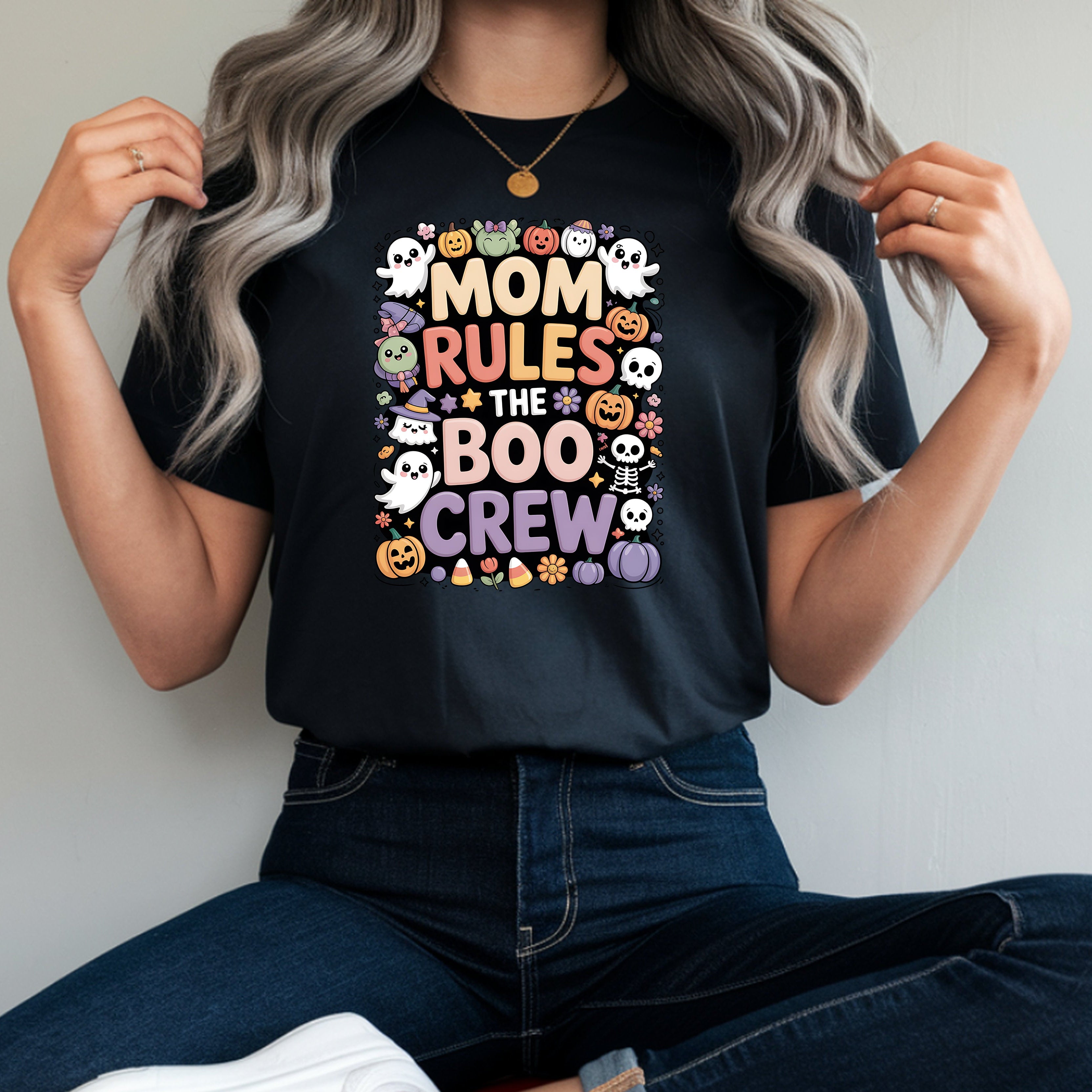 A woman wearing a black graphic t-shirt with a colorful 'Mom Rules the Boo Crew' Halloween design, paired with dark blue jeans and white sneakers, sitting on a red stool.