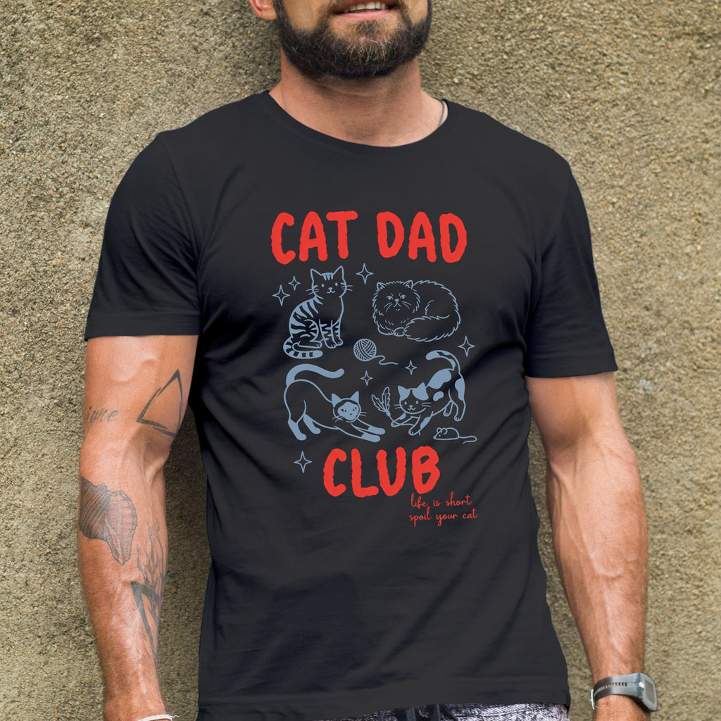 Man wearing a black t-shirt with 'Cat Dad Club' text and graphics against a textured wall.