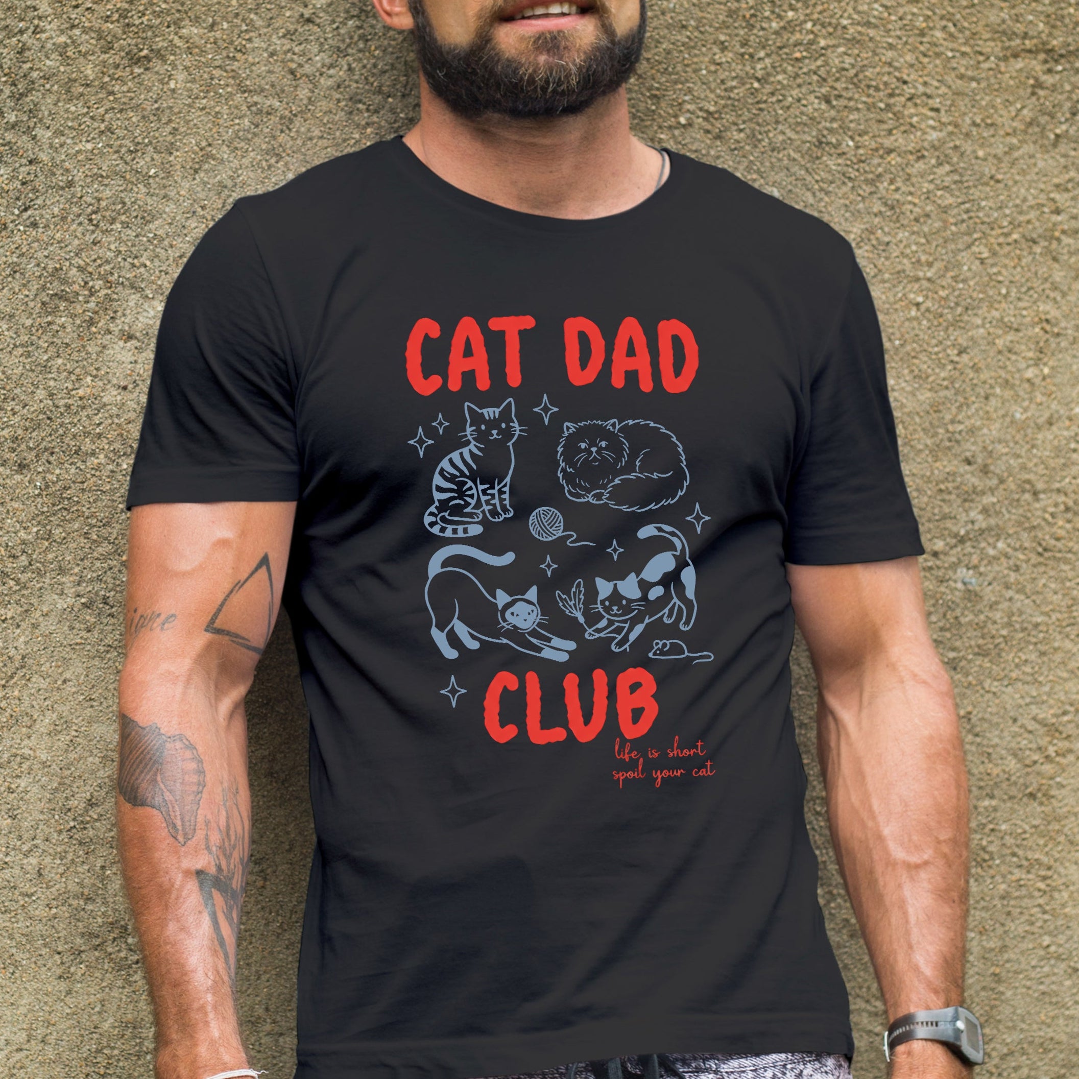 Man wearing a black t-shirt with 'Cat Dad Club' text and graphics against a textured wall.
