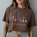 A woman wearing a brown cropped t-shirt with a graphic of colorful witch brooms and the text 'A girl gotta have options'.