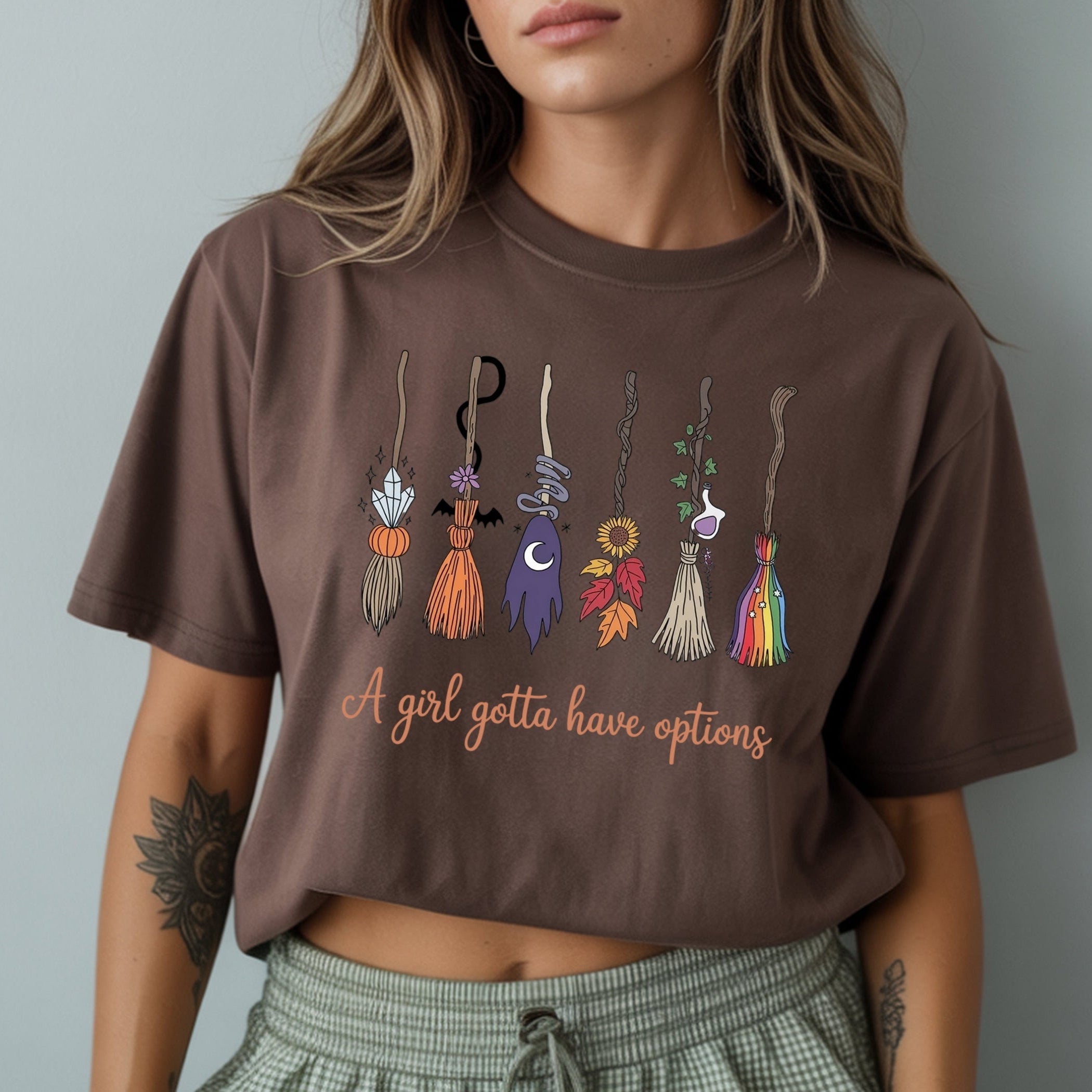 A woman wearing a brown cropped t-shirt with a graphic of colorful witch brooms and the text 'A girl gotta have options'.
