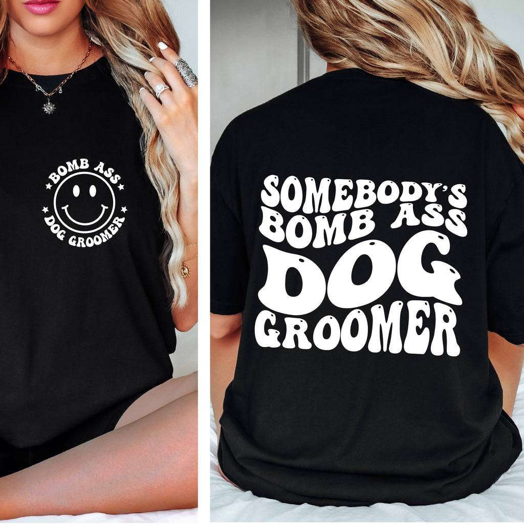 Black t-shirt with 'Somebody's Bomb Ass Dog Groomer' text and smiley face design on a person.