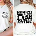 White t-shirt with 'Bomb Ass Lash Artist' text and smiley face on front, worn by a person.