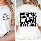 White t-shirt with 'Bomb Ass Lash Artist' text and smiley face on front, worn by a person.