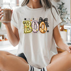 A woman sitting cross-legged on a kitchen counter wearing a white graphic t-shirt with a colorful 'BOO' donut design, holding a mason jar of iced coffee.