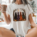 A woman sitting cross-legged in a bright kitchen wearing a white graphic t-shirt with a Halloween witch illustration, holding an iced coffee.