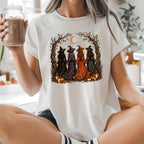 A woman sitting cross-legged in a bright kitchen wearing a white graphic t-shirt with a Halloween witch illustration, holding an iced coffee.