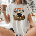 A woman sitting cross-legged in a bright kitchen wearing a white graphic t-shirt that reads 'Spooky Biscuits Bakery' with a black cat illustration. She is holding a mason jar filled with a coffee drink.