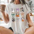 A woman sitting cross-legged in a bright kitchen wearing a white graphic t-shirt with a Halloween design featuring cartoon geese, pumpkins, and bows. She is holding a glass jar of iced coffee.