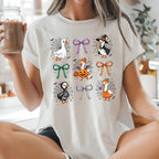 A woman sitting cross-legged in a bright kitchen wearing a white graphic t-shirt with a Halloween design featuring cartoon geese, pumpkins, and bows. She is holding a glass jar of iced coffee.