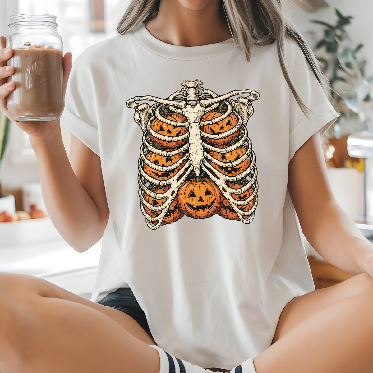A woman sitting cross-legged on a kitchen counter wearing a white graphic t-shirt with a Halloween ribcage design made of jack-o'-lanterns. She is holding a glass of iced coffee in a bright, modern kitchen.