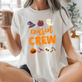 A woman sitting cross-legged in a bright kitchen wearing a white graphic T-shirt that says 'cousin CREW' in orange with Halloween-themed illustrations. She is holding an iced coffee in a mason jar.