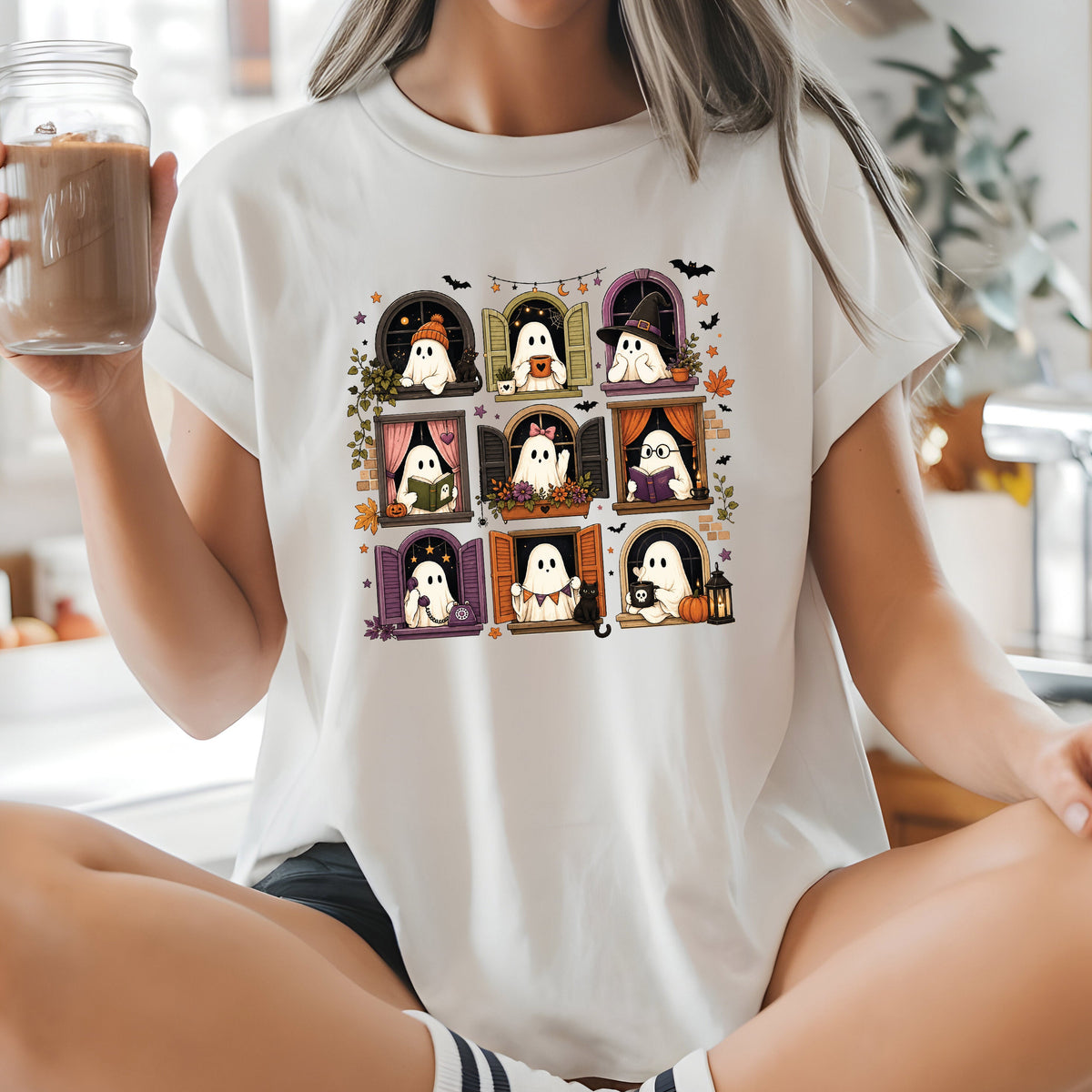 A woman sitting cross-legged on a kitchen counter wearing a white graphic t-shirt with a 3x3 grid of cute cartoon ghosts in Halloween-themed frames. She is holding a glass jar of iced coffee in a bright, modern kitchen.