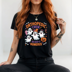 A woman wearing a black graphic t-shirt with a Halloween orthopedic theme. The shirt features cartoon ghosts, a witch hat, a jack-o'-lantern, and the text 'ORTHOPODIC CREW WE KEEP IT HUMERUS'.