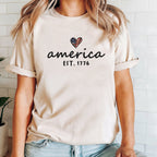 Person wearing a beige t-shirt with 'america est. 1776' text and heart design.