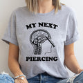 Person wearing a gray t-shirt with a graphic design and text 'My Next Piercing' on a plain background
