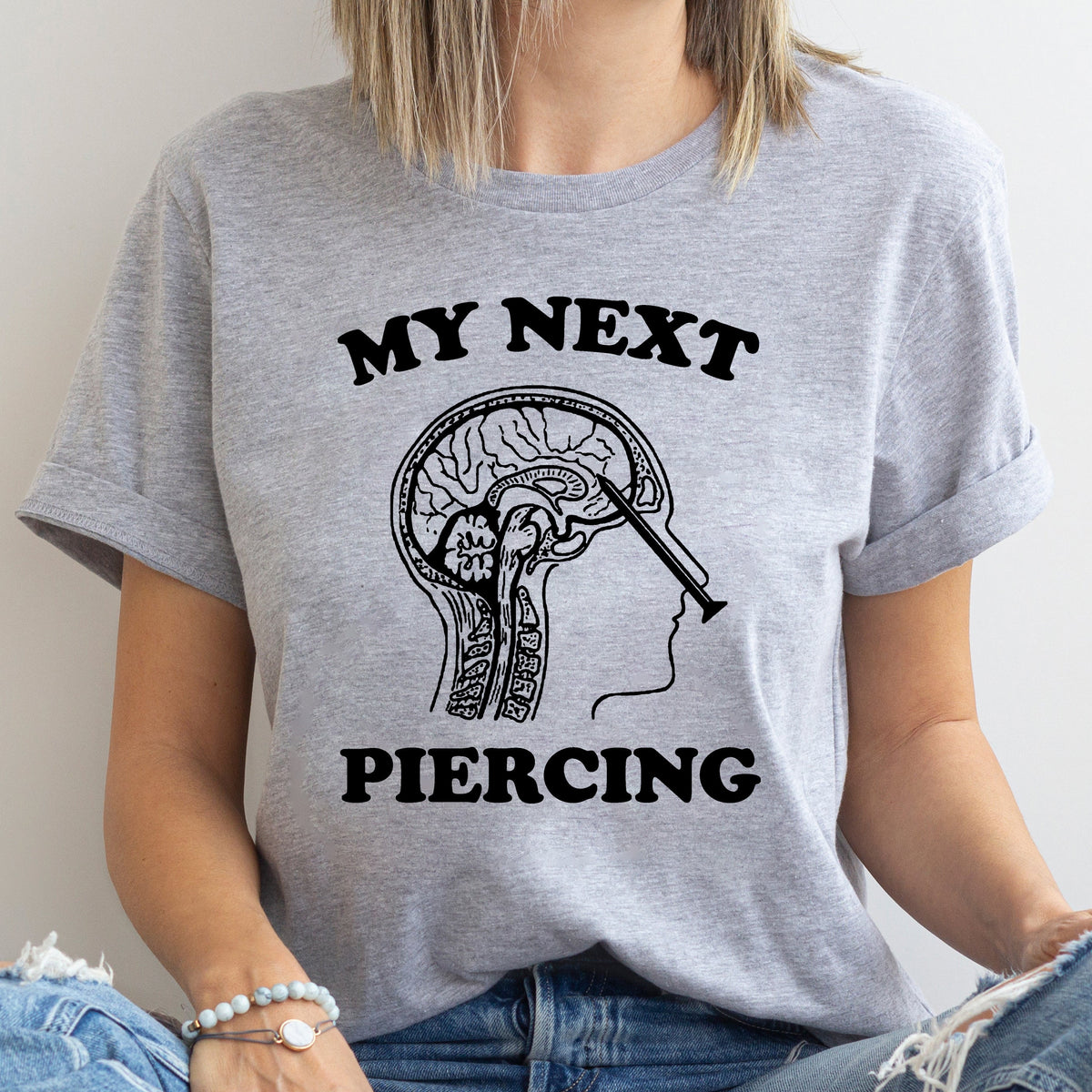 Person wearing a gray t-shirt with a graphic design and text 'My Next Piercing' on a plain background