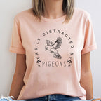 Person wearing a peach t-shirt with a graphic and text 'Easily Distracted by Pigeons' on a light background