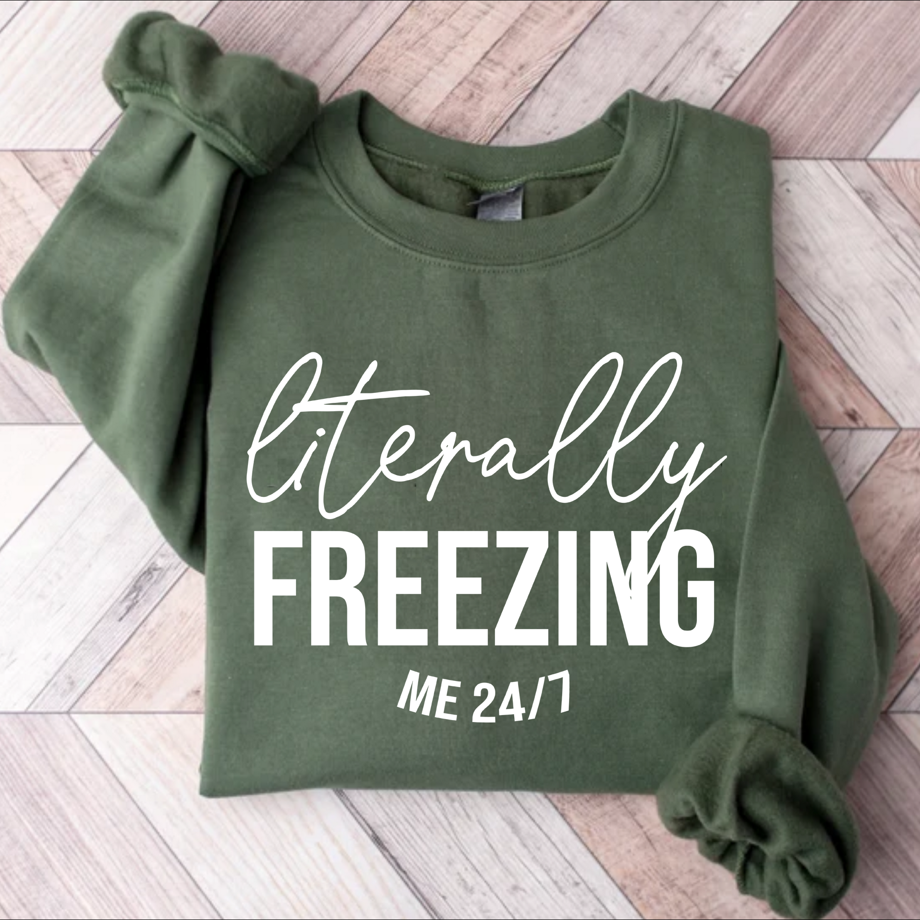 Green sweatshirt with 'Literally Freezing Me 24/7' text on a wooden floor.