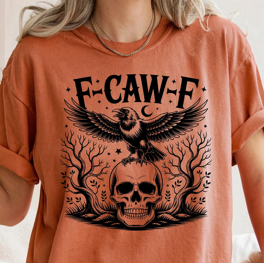 A person wearing a burnt orange t-shirt with a black graphic print featuring the text 'F-CAW-F', a crow, a skull, and tree branches.