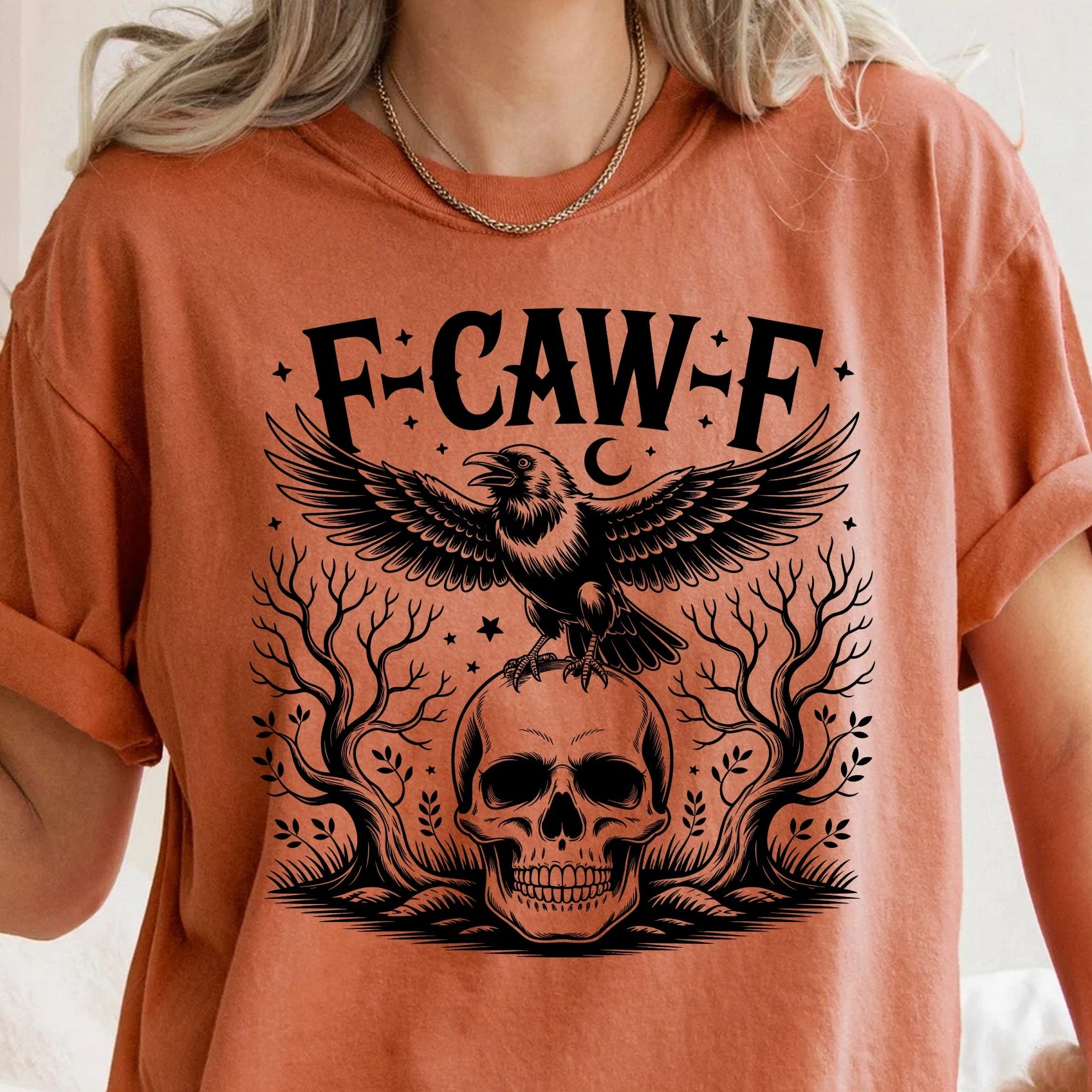 A person wearing a burnt orange t-shirt with a black graphic print featuring the text 'F-CAW-F', a crow, a skull, and tree branches.